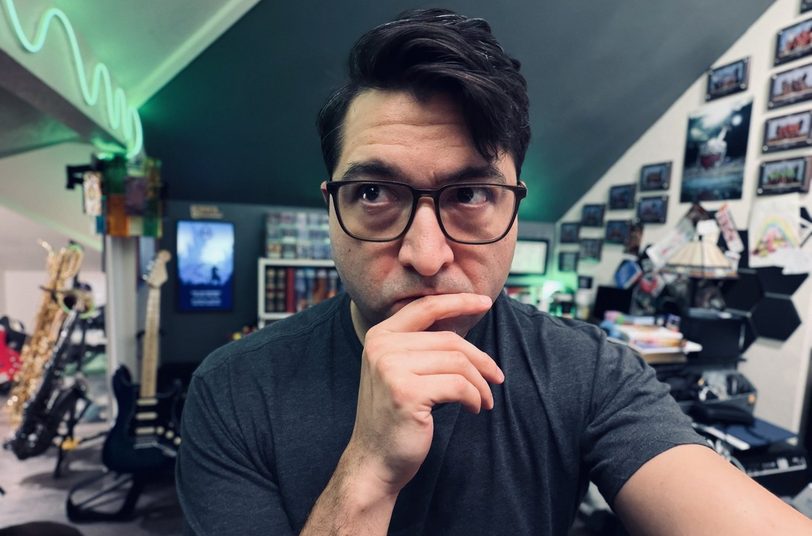 Tim, in glasses and a grey t-shirt, hand on chin, in a studio with guitars and saxophones.